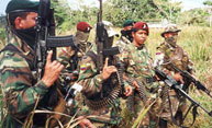 A small group of men outfitted in camouflage military uniforms wearing berets, some red and some black, stand in tall grass in a jungle clearing holding AK-47 rifles.